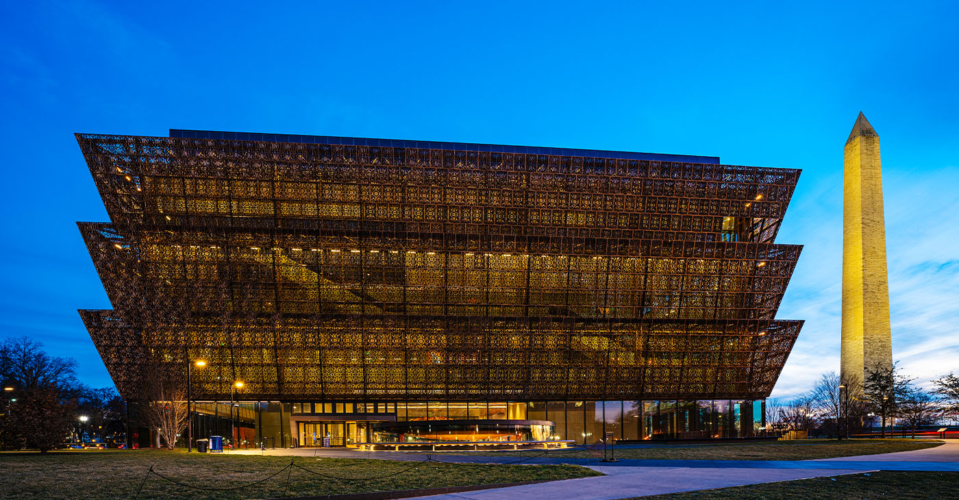 A photo of the National Museum of African American History and Culture at night with the Washington Monument in downtown Washington DC.